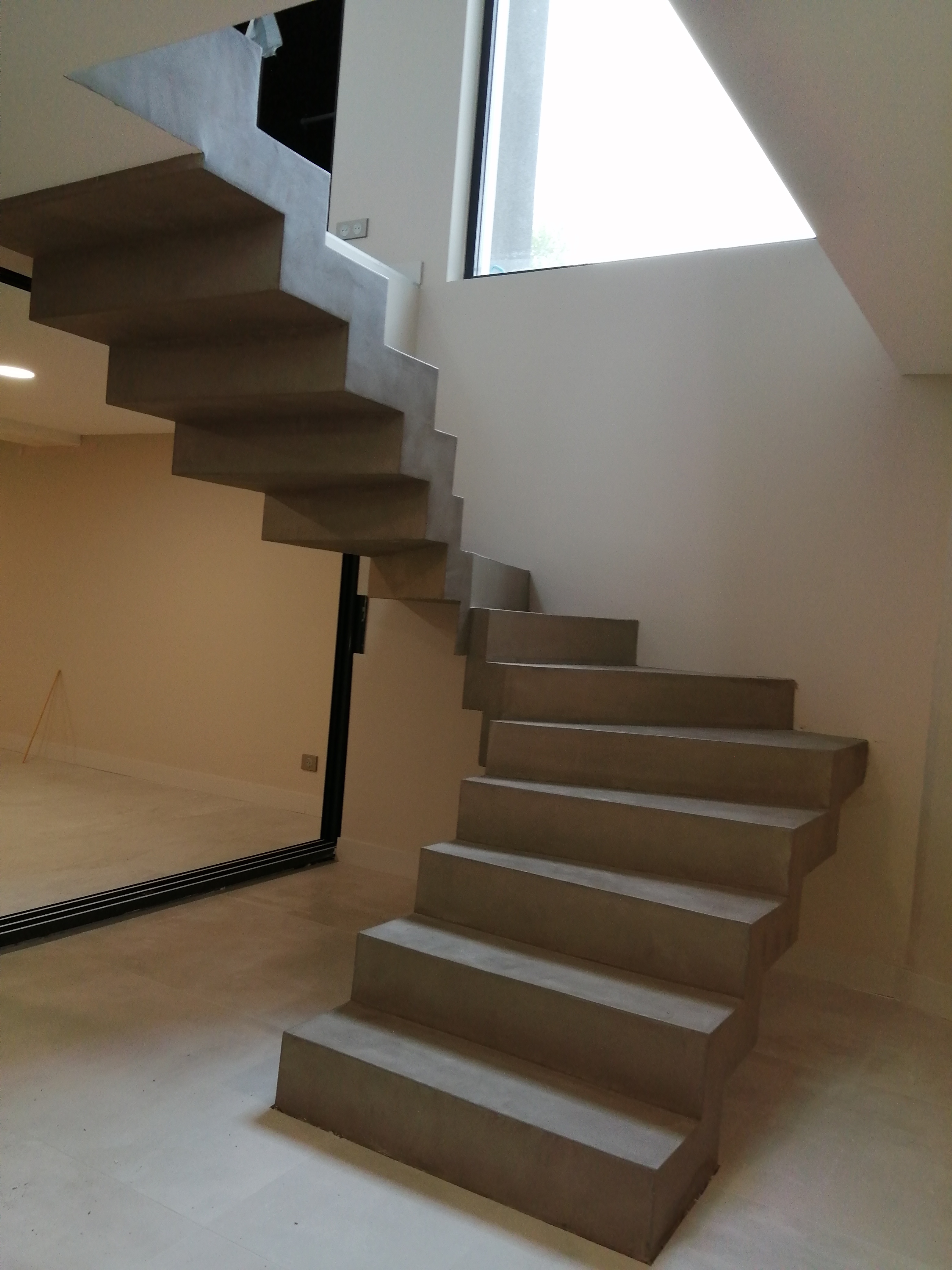 Finition béton ciré Persan sur un fond beige clair au mur illuminé par une baie vitrée sur toute la hauteur de la cage d'escalier